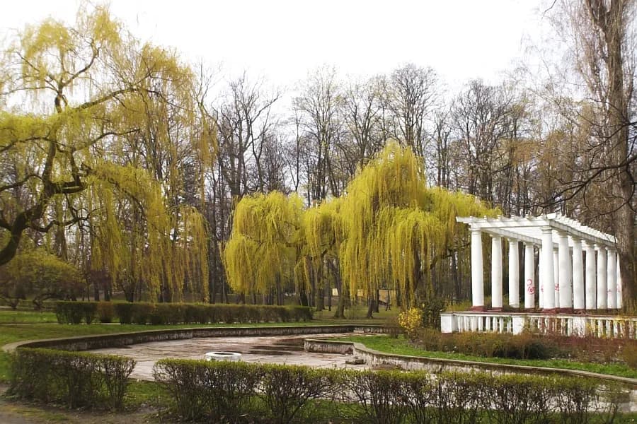 A park scene features weeping willow trees with bright yellow leaves, a white classical colonnade, and an empty, ornate pond or fountain basin with surrounding hedges and grassy paths.